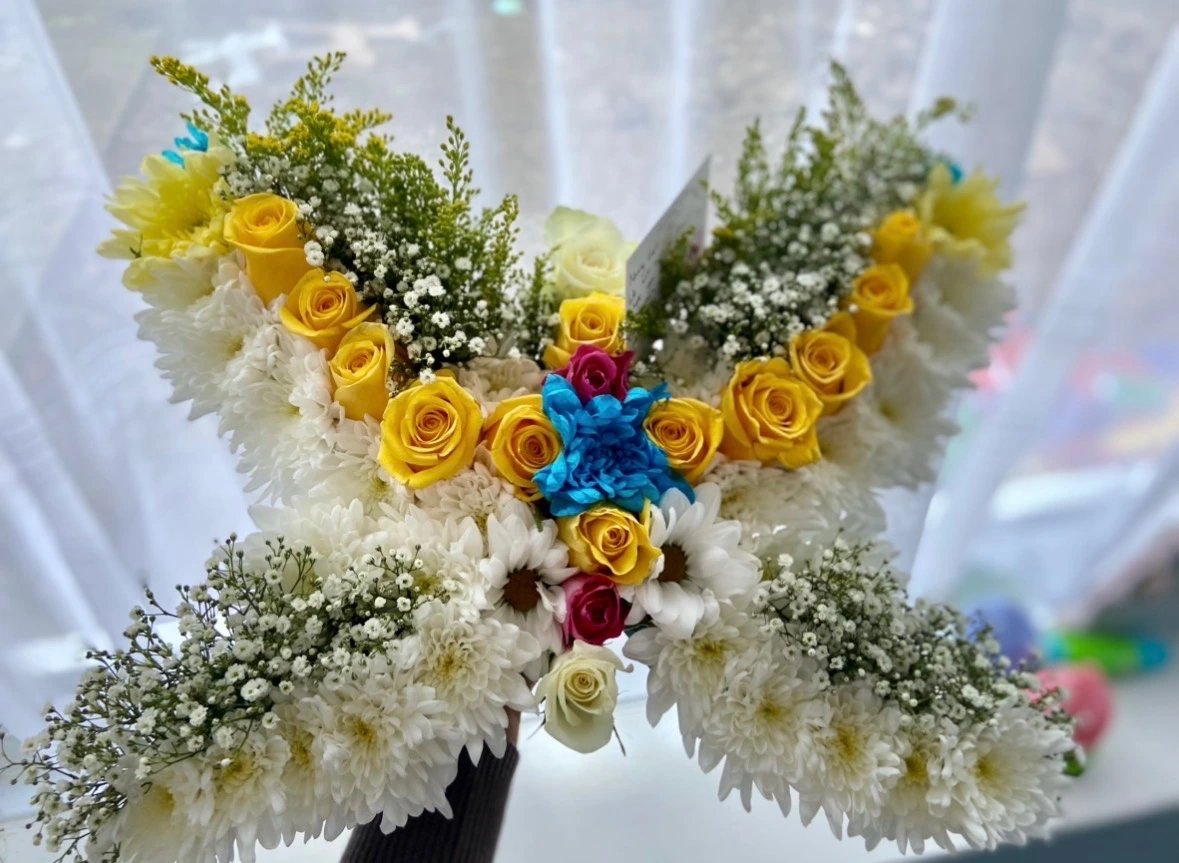 A fresh butterfly tribute in white chrysanthemums with yellow roses, blue and pink accent blooms, and gypsophila