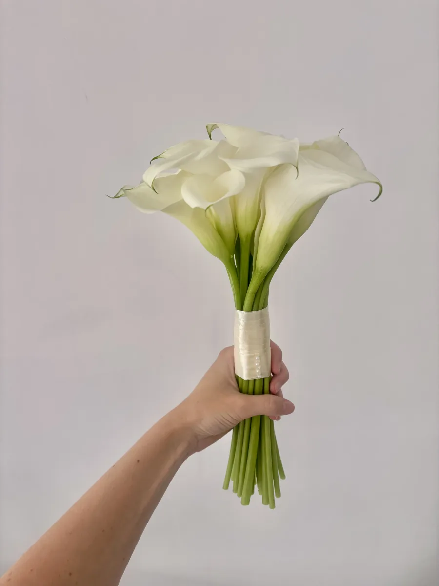 A fresh bridal bouquet of white calla lilies bound with ivory satin ribbon, held by the bride