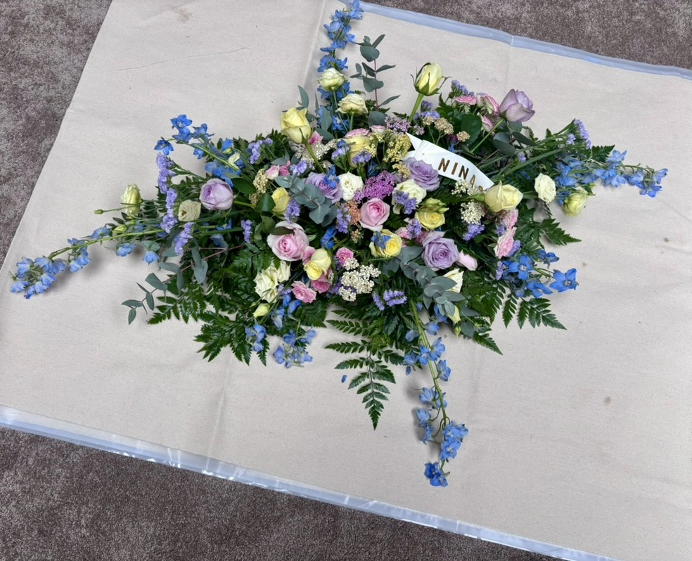 A pastel fresh funeral spray of lilac, pink, and yellow roses with blue delphinium and trailing eucalyptus